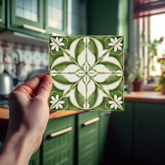 a hand holding up a green and white tile in a kitchen with green cupboards