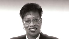 a black and white photo of a woman wearing glasses, a suit and tie smiling for the camera