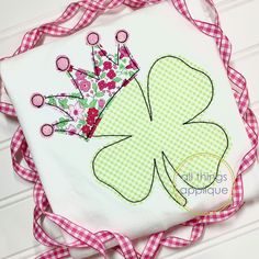 a green and white shamrock with a crown on it's head is sitting on a pink checkered ribbon