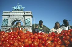 several people standing around a pile of tomatoes in front of an arch with statues on top