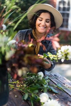 a woman in a hat smiles as she arranges flowers on a wooden table outside