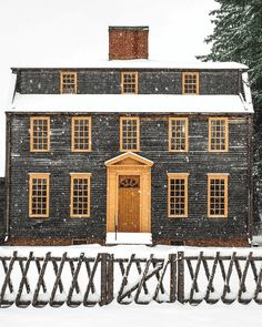 a black house covered in snow next to a wooden fence and evergreen trees on a snowy day