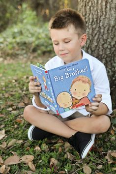 a young boy sitting on the ground reading a book in front of a tree with leaves around him