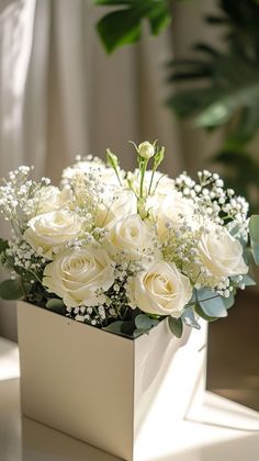 white roses and baby's breath in a square vase on a table with greenery