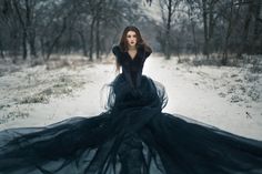 a woman in a long black dress sitting on the snow covered ground with trees behind her