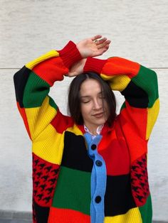 a woman wearing a multi colored sweater and holding her hands up to her head while standing in front of a white wall