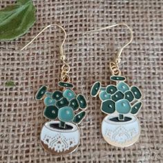 two green and white earrings sitting on top of a piece of cloth next to a plant