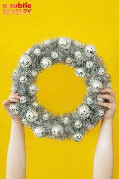 two hands holding up a wreath with silver ornaments on it, against a yellow background