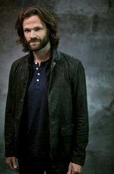 a man with long hair and beard standing in front of a dark background wearing a leather jacket