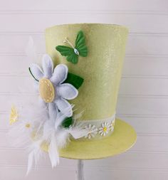 a yellow top hat with white flowers and green leaves on the side, sitting on a stand