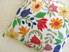 a white pillow with multicolored flowers on it sitting on a tablecloth covered surface