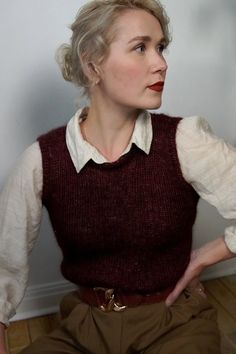 a woman sitting on the floor with her hands in her pockets and wearing a sweater vest