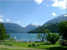 a lake surrounded by mountains and trees with people on the grass in front of it