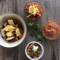 three bowls filled with chili, cheese and nachos on top of a wooden table