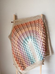 a crocheted blanket is hanging on a wooden stand with a white wall in the background