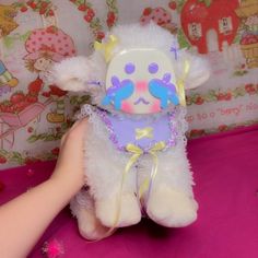 a white stuffed animal with purple and yellow decorations on it's face, being held by a hand