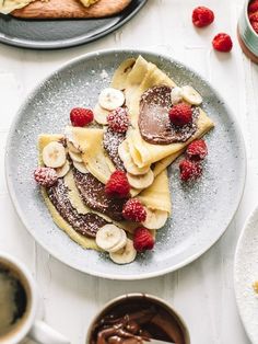 pancakes with bananas, raspberries and chocolate sauce on a plate next to two cups of coffee