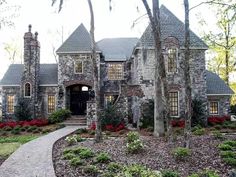 a stone house surrounded by trees and flowers in the front yard with a walkway leading to it