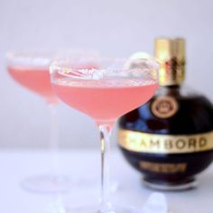 two glasses filled with pink liquid next to a bottle of alcohol on a white table