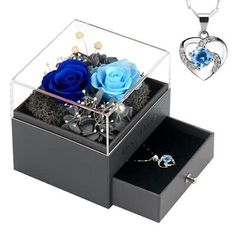 two blue roses are in a clear box and one has a heart shaped pendant on it