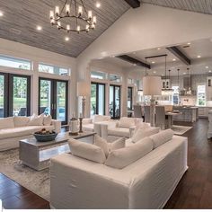 a living room filled with white couches and chairs next to a kitchen area covered in windows