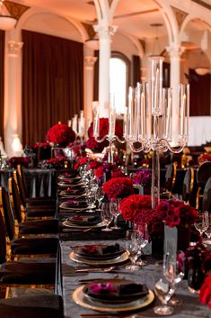 a long table is set with black and gold place settings, red flower centerpieces, and silverware