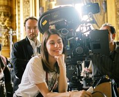 a woman sitting in front of a camera on top of a wooden table next to other people