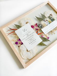 the wedding stationery is in a wooden frame with flowers and leaves on it's side