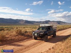 a pick up truck driving down a dirt road in the desert with mountains in the background