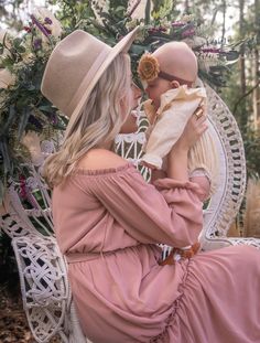 a woman in a pink dress and hat sitting on a white wicker chair holding a baby