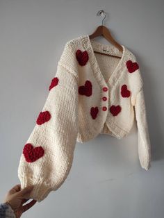 someone holding up a knitted sweater with hearts on the front and back, hanging from a hook