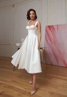 a woman in a white dress standing next to a painting on a wooden floor and posing for the camera