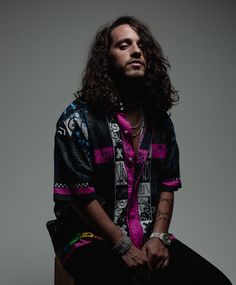 a man with long hair sitting on top of a wooden table wearing a shirt and pants