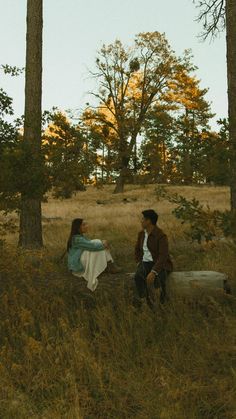 a man and woman sitting on a log in the middle of a field with trees