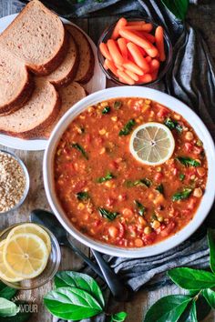 a white bowl filled with soup next to sliced bread, carrots and lemon slices