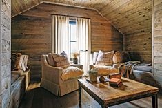 a living room with wooden walls and flooring in the corner is furnished with two couches, a coffee table, and a window