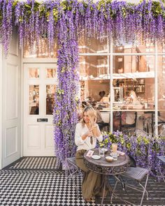 a woman sitting at a table in front of a building with purple flowers on it