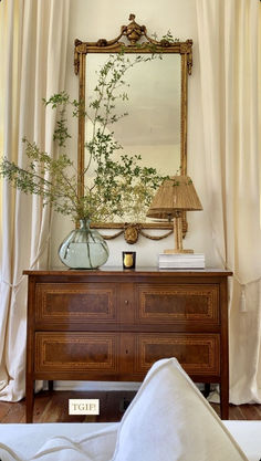 a dresser with a mirror, lamp and flowers on it in front of a white curtained window