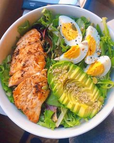 a white bowl filled with chicken, avocado and hard boiled eggs on top of lettuce