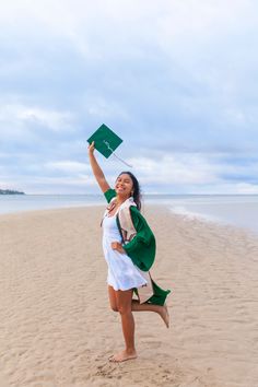 a woman in white dress and green graduation cap holding up a green flag on the beach