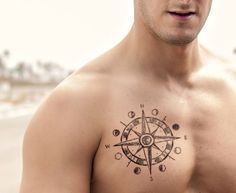 a shirtless man with a compass tattoo on his chest looking at the camera while standing in front of an ocean