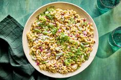 a white bowl filled with pasta salad on top of a green cloth next to two glasses