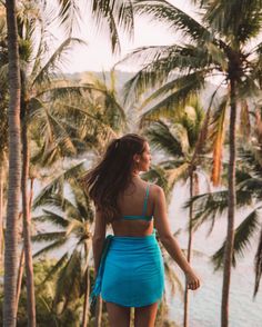 a woman in a blue dress is walking by the water with palm trees behind her