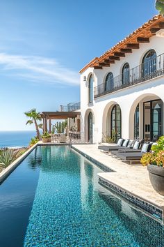 an outdoor swimming pool in front of a large house overlooking the ocean with chaise lounges