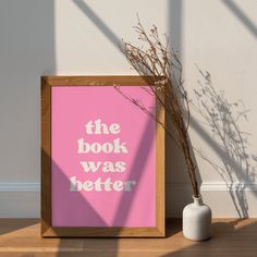 a pink poster with the words the book was better next to a white vase on a wooden floor