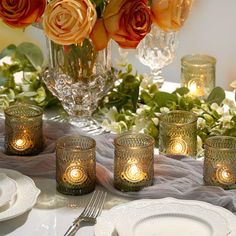 there are many candles on the table with flowers in the vase behind them and plates around it