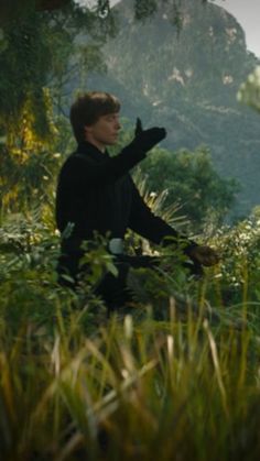 a young man sitting in the grass with his hands out and pointing to something behind him