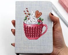 a hand holding a cross stitch book with a cup of hot chocolate and marshmallows in it