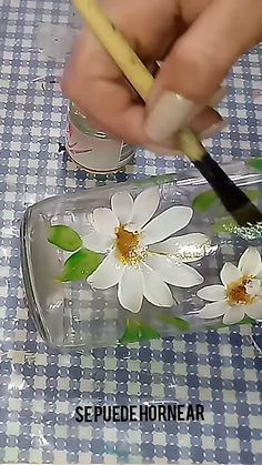 a person is holding a pencil and painting flowers on a clear glass container with a yellow handle