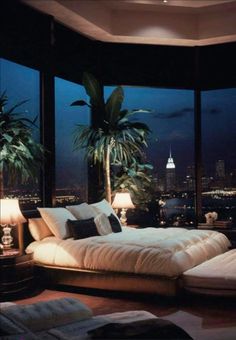 a large bed sitting in the middle of a bedroom next to two windows with city lights on them